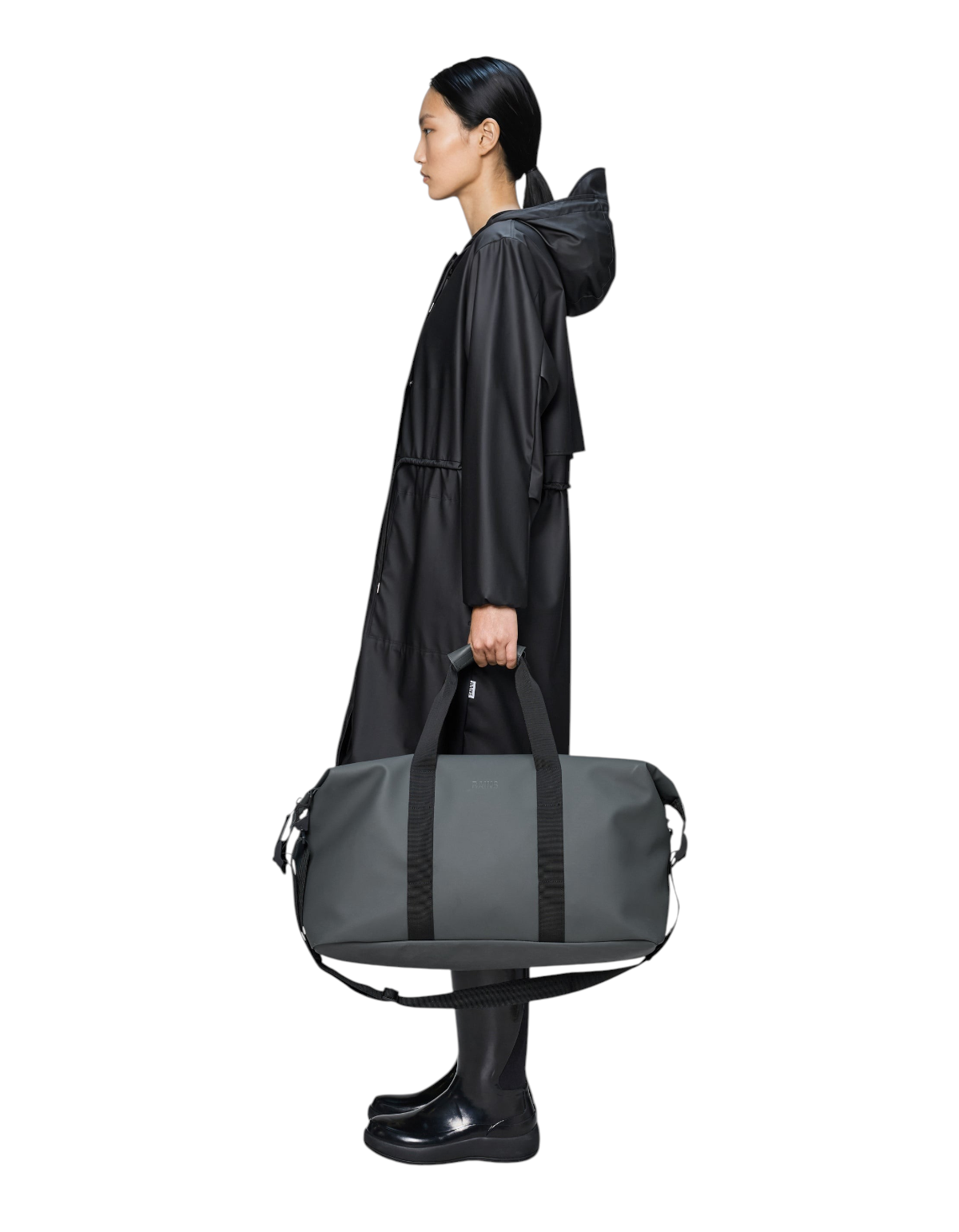 Modella che indossa un impermeabile nero mentre tiene il borsone DevidLabel Rains Hilo Weekend Bag grigio scuro
