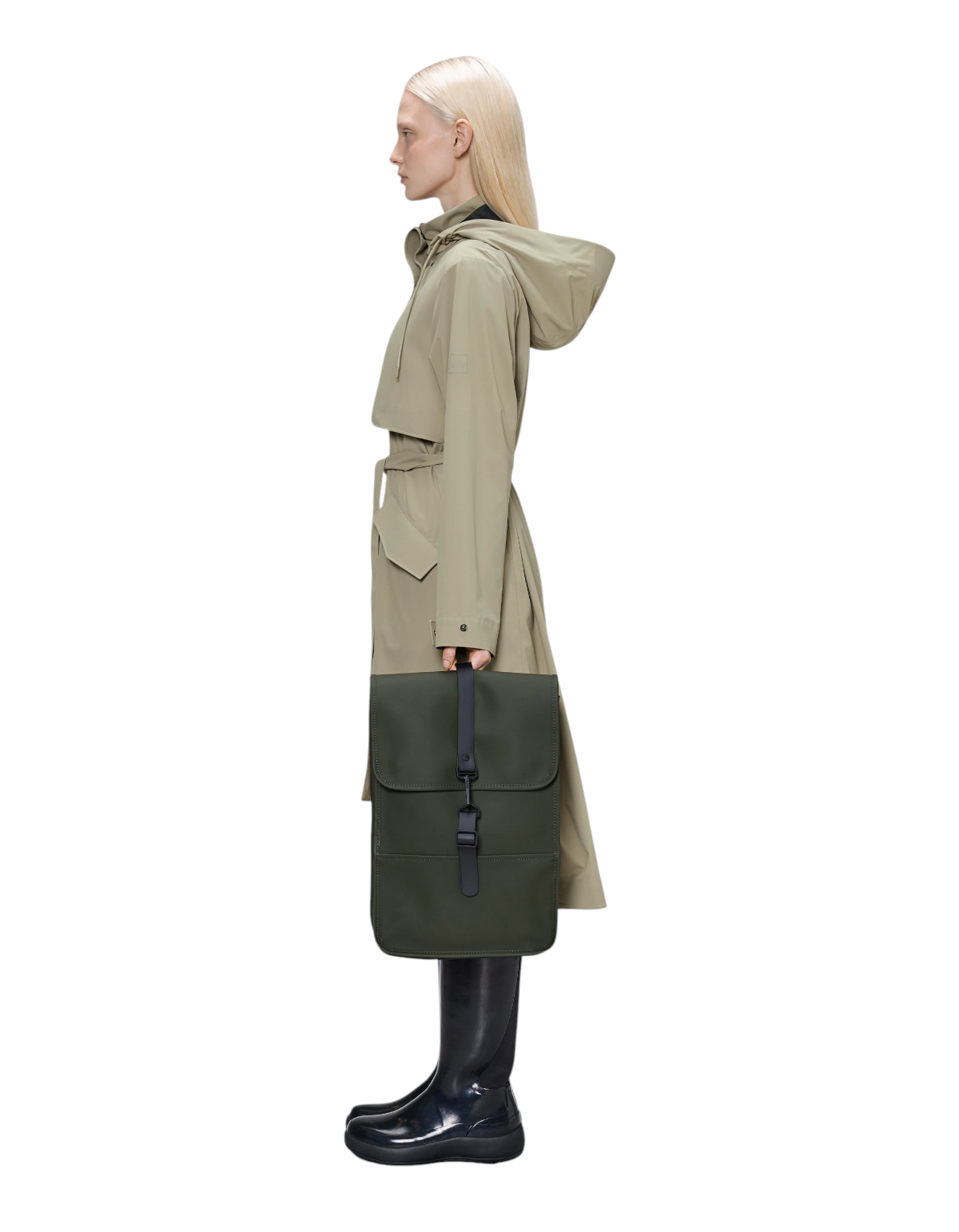 Modella di profilo indossa un impermeabile beige e tiene in mano lo zaino DevidLabel Rains Backpack Mini verde