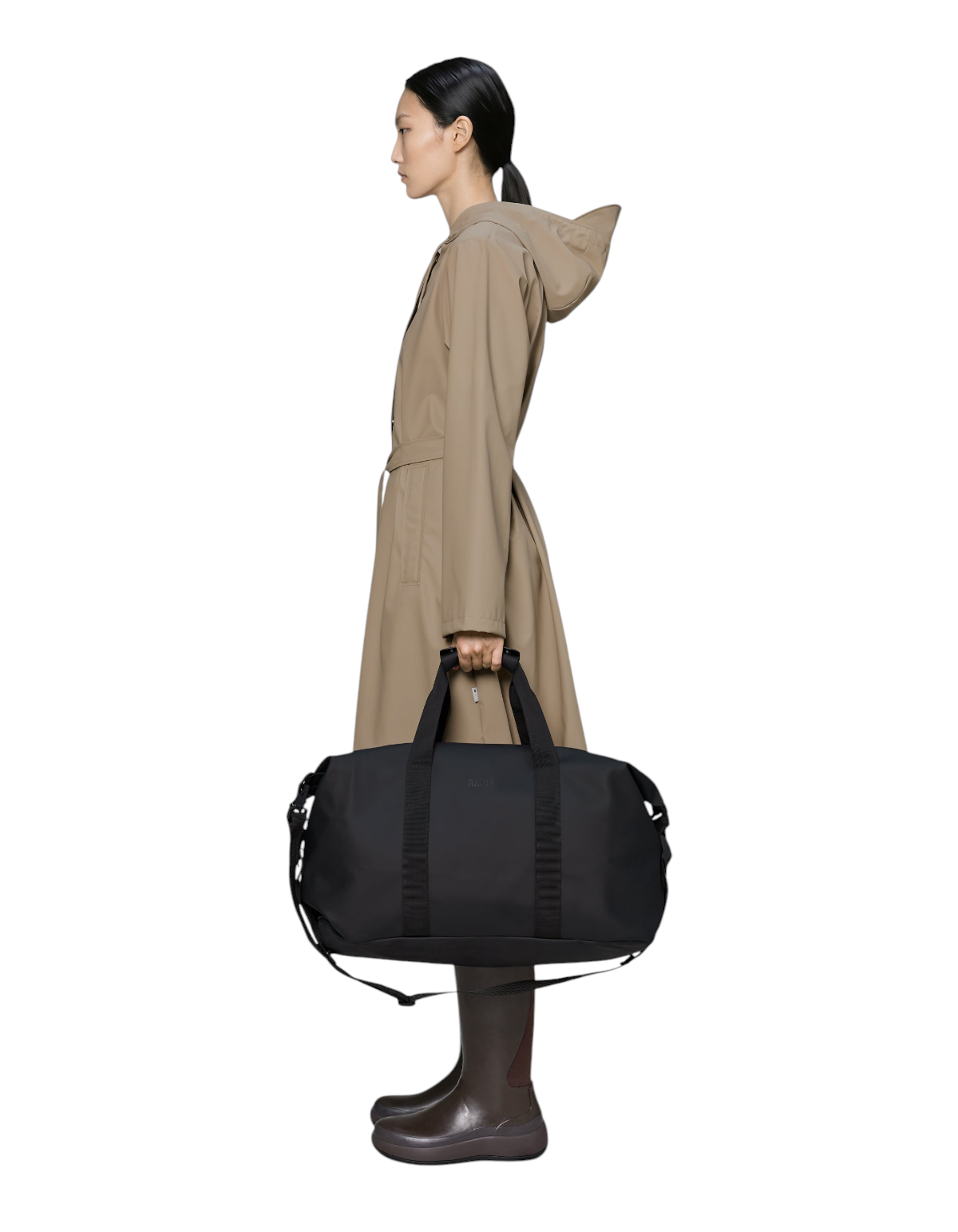 Modella che indossa un impermeabile beige mentre tiene in mano il borsone nero DevidLabel Rains Hilo Weekend Bag