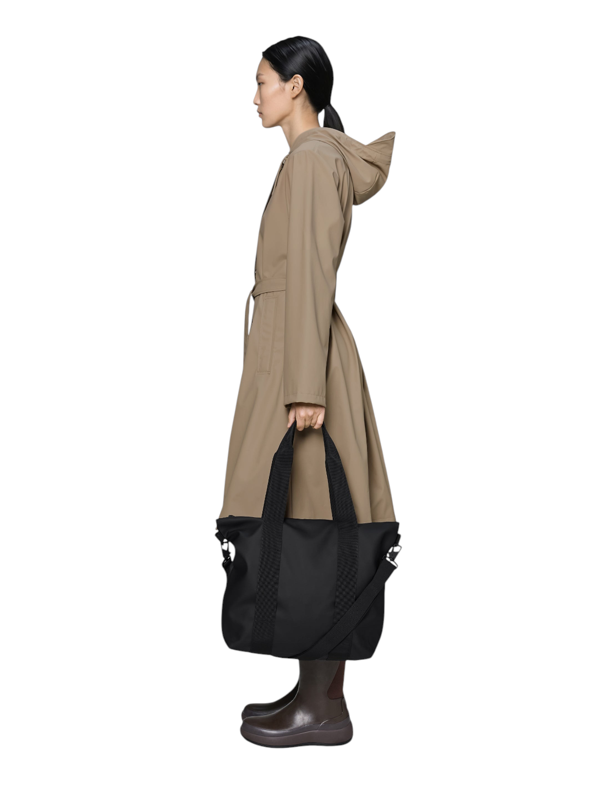 Modella di profilo indossa un impermeabile beige e tiene in mano la borsa nera Rains Tote Bag Mini di DevidLabel