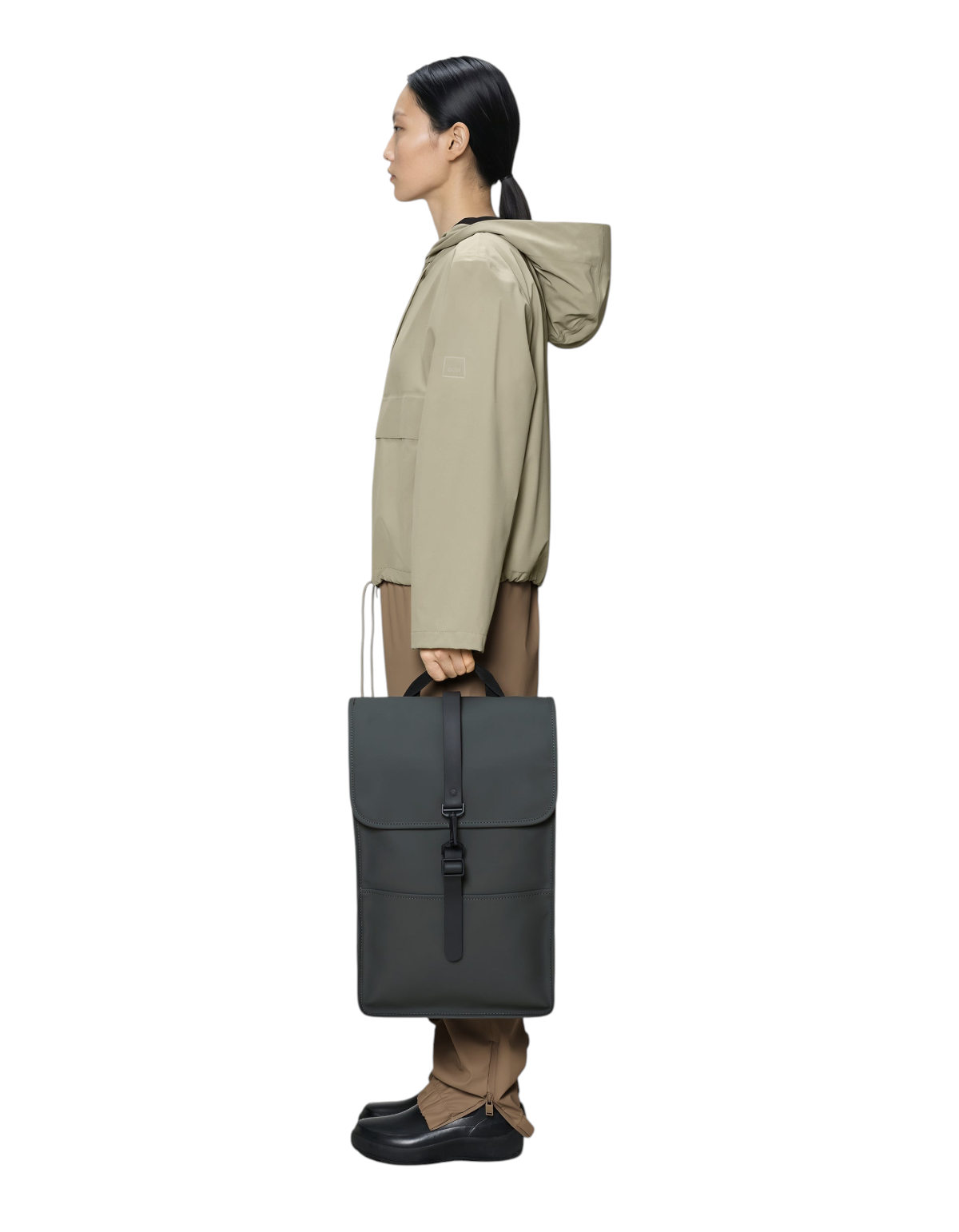 Modella di profilo tiene lo zaino impermeabile Rains Backpack W3 in grigio scuro, design minimalista e moderno.